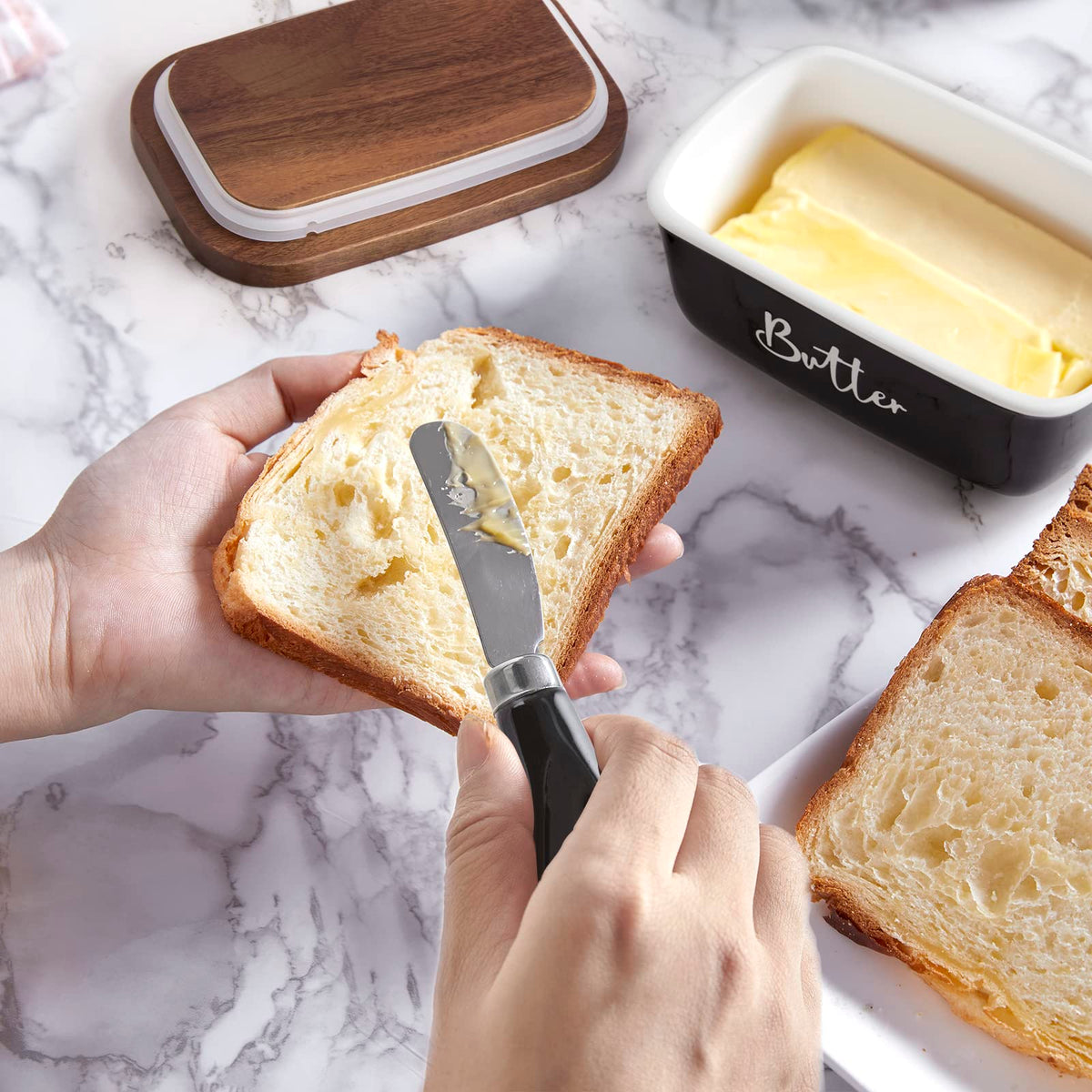 Hands spreading butter on bread beside the black butter dish