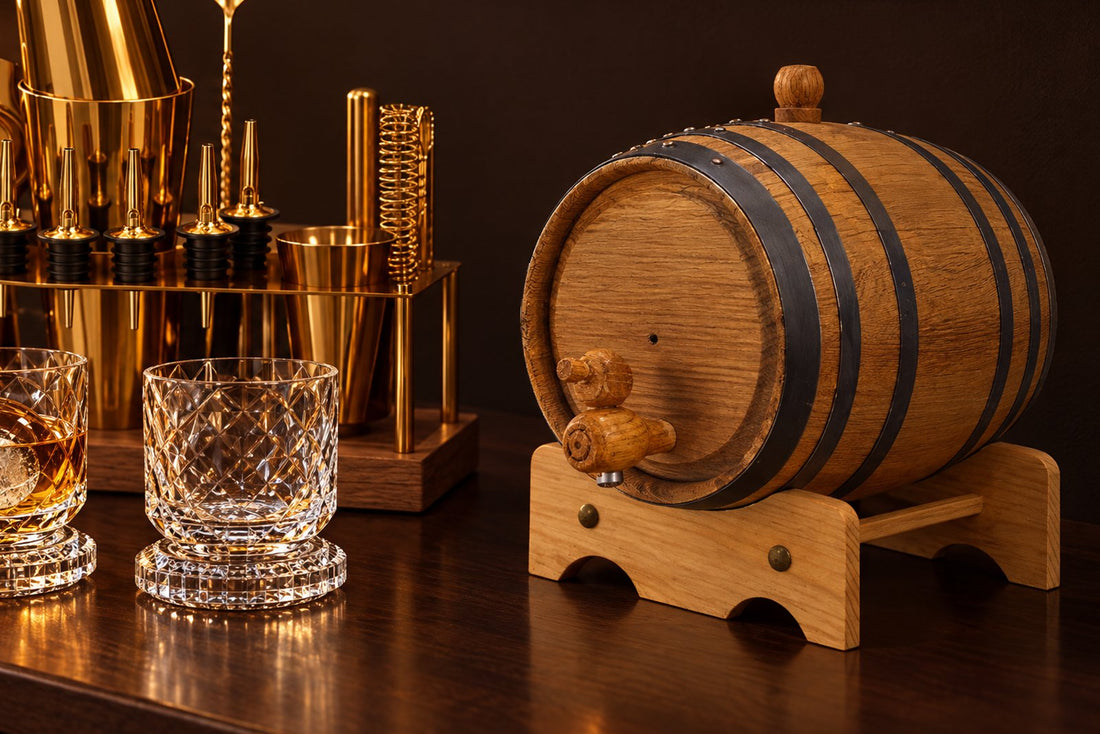 Small charred oak barrel with a wooden spigot on its stand beside diamond-cut whiskey glasses and a gold cocktail set on a walnut bar